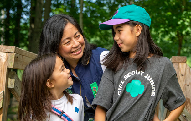 A smiling adult Girl Scout volunteer and two smiling youth members wearing Girl Scout apparel stand outdoors. There are trees in the background.