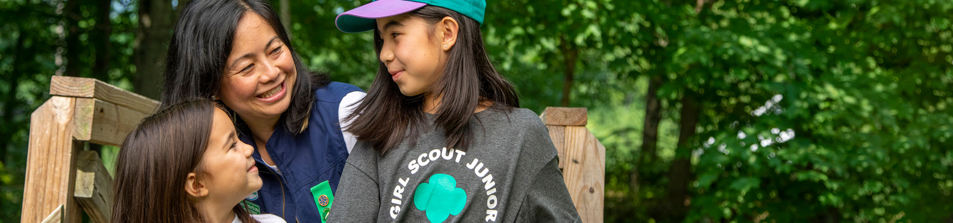  Adult Girl Scout volunteer and two youth Girl Scout members wearing Girl Scout apparel stand outdoors with trees behind them. 