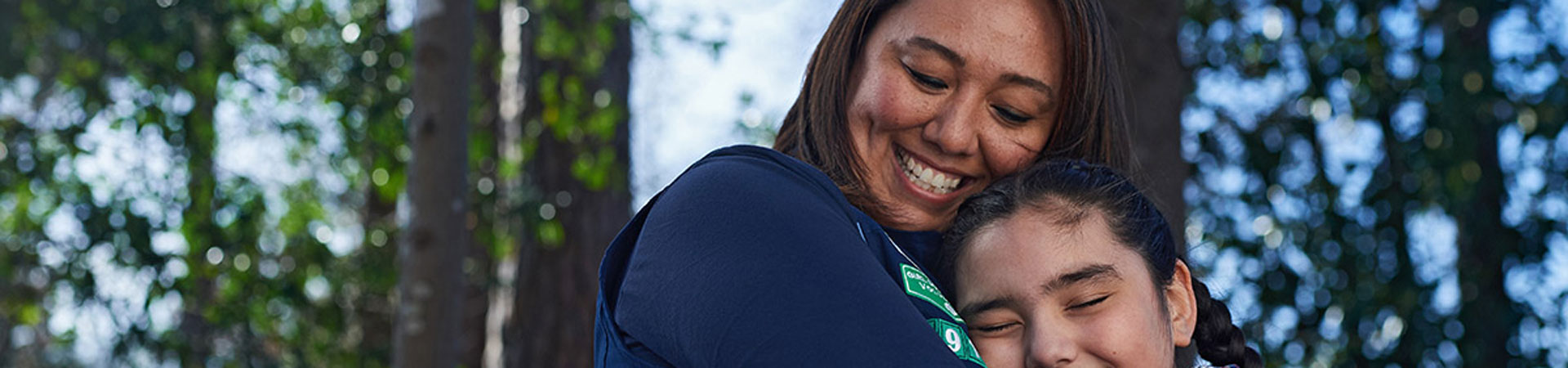  An adult volunteer in a blue vest hugging a youth Girl Scout member. They are outdoors and there are trees behind them.  