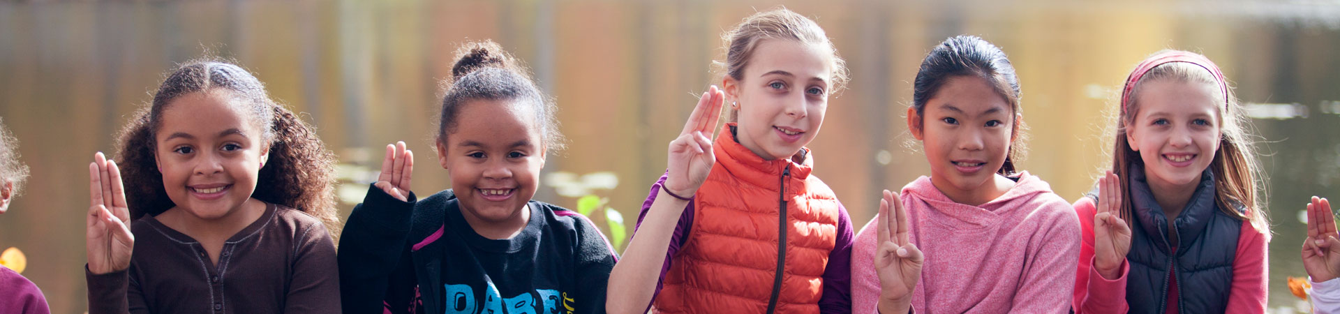  Five youth Girl Scouts sit in a row outdoors. They are smiling and making the Girl Scout sign with their hands. 