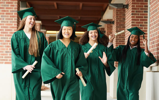 Four Girl Scout alums dressed in green graduation caps and gowns are holding diplomas and walking down a brick corridor.