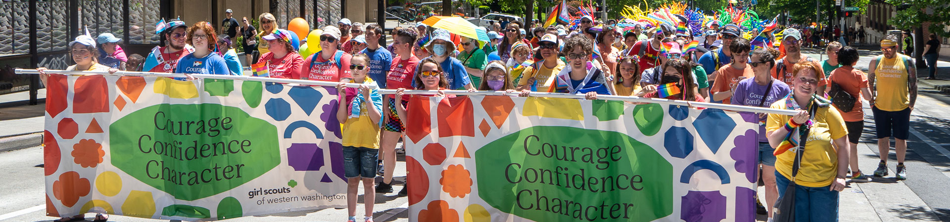  Girl Scout staff, youth, and adult members march in the Seattle Pride parade. It is sunny, and they are wearing colorful clothing and waving rainbow flag. The people at the front are holding a large, rainbow-colored banner that reads, "Courage, Confidence, Character." 