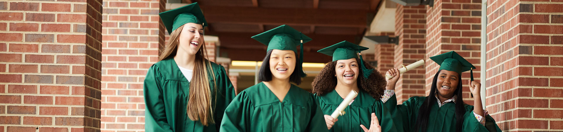  A group of Girl Scouts dressed in green graduation caps and gowns walk down a brick corridor with diplomas in their hands. 