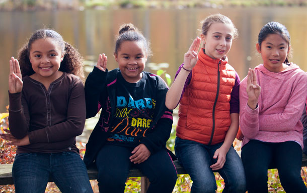 Four Girl Scouts sit on a bench in front of a body of water, making the Girl Scout Promise sign.