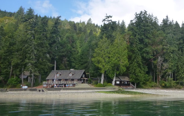 A camp lodge sits on the edge of a rocky beach with trees in the background.