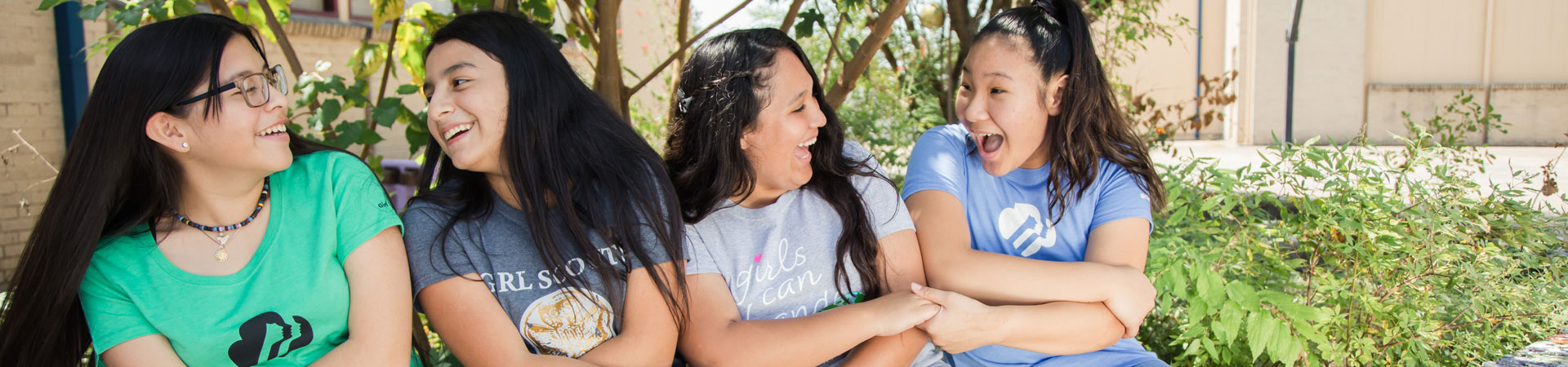  Four Girl Scout alum are sitting outdoors, smiling, with their arms linked. 