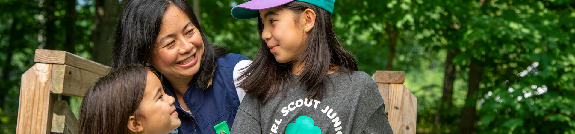  Two smiling youth Girl Scouts and a caregiver are outside next to a wooden bench with trees behind them. 