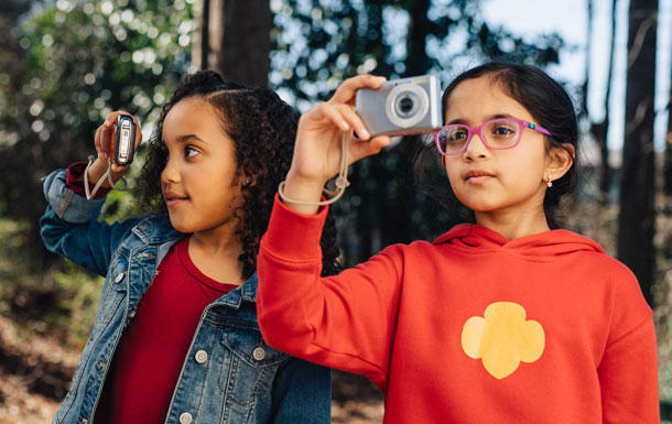 A Girl Scout in an orange sweatshirt with a yellow Trefoil holds a camera while standing next to a Girl Scout in a red shirt with a jean jacket.