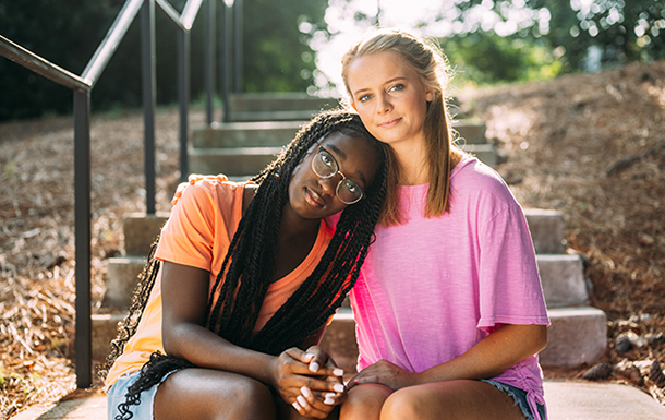 Two Girl Scouts sitting on steps outdoors. One Girl Scout is resting their head on the shoulder of the other consoling Girl Scout.