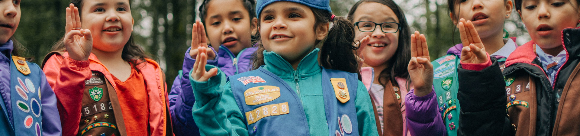  A group of younger Girl Scouts making the Girl Scout sign stand outside surrounded by trees. They are dressed in badge-decorated uniforms. 