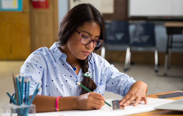 A woman with glasses sitting at a desk writing with a pencil. 