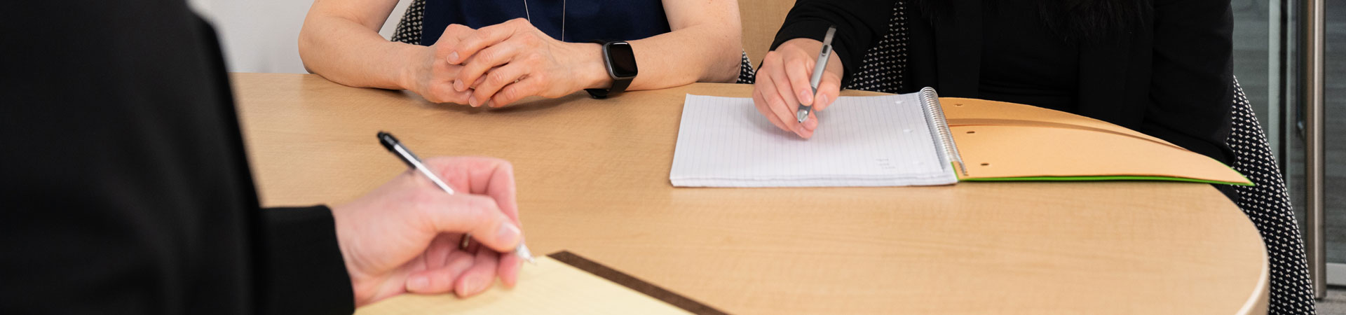  The torsos and arms of three people seated around a wooden table. Two of the people are taking notes. 