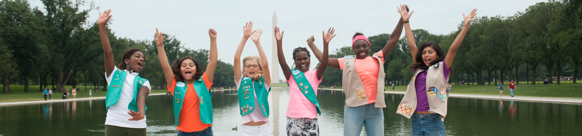  Six Girl Scouts wearing Girl Scout vests stand in a row with their arms raised overhead. Behind them is the Lincoln Memorial Reflecting Pool and Washington Monument and the background. 
