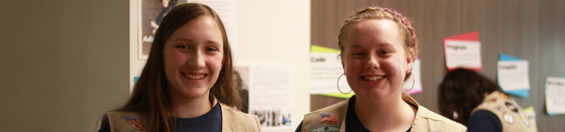  Two Girl Scouts wearing tan vests and dark blue shirts that read Global Action Team in white letters stand together and smiling in front of brown wood cabinets. 