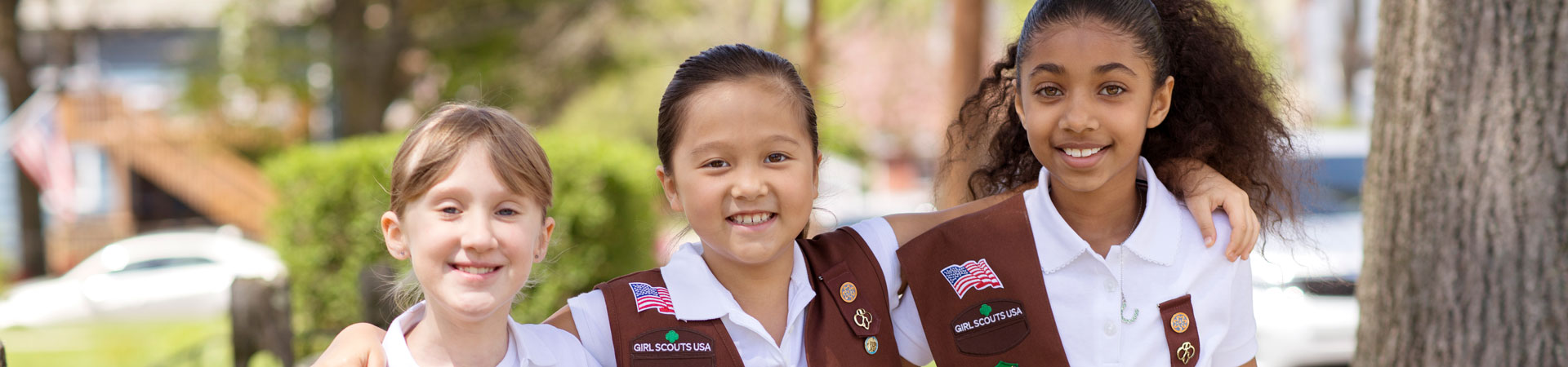  Three Brownies wearing brown vests and sashes stand arms around each other smiling outside on a sidewalk. 