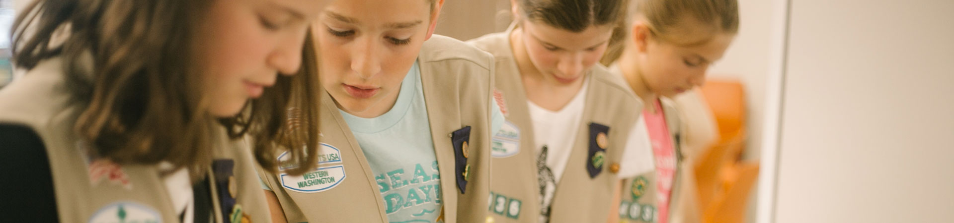  Four Girl Scouts wearing tan vests are seen standing at a lab table, working on a science experiment that involves connecting red, green, yellow, and white wires. 