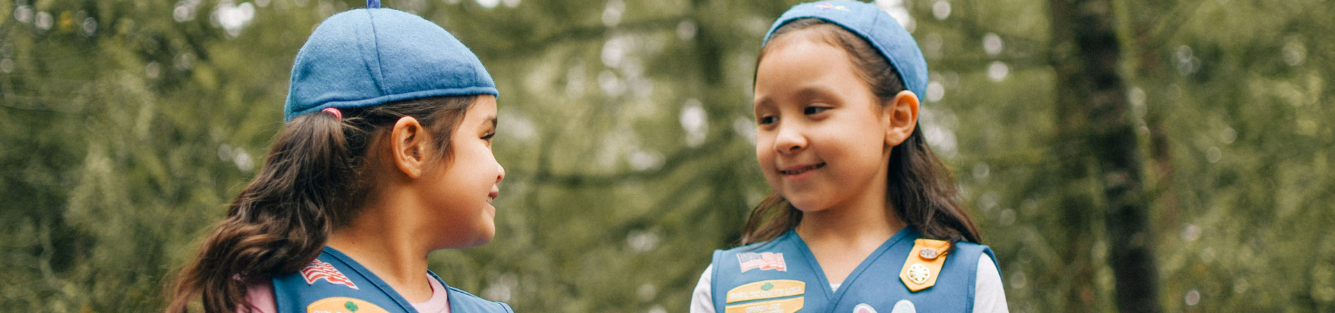  Two Daisies wearing blue vests and hats smile at each other outside in front of green trees. 