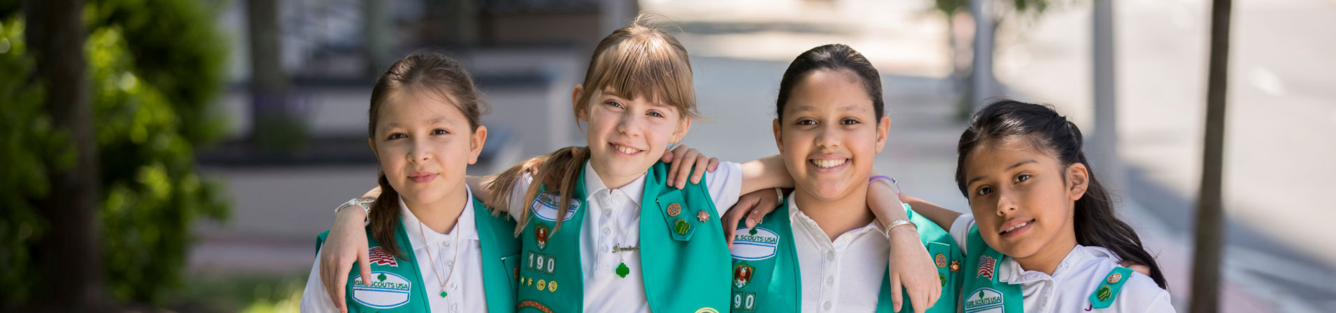  Four Girl Scouts wearing white shirts with green vests and khaki pants and skirts are standing outside on a sidewalk, arms around each other and smiling. 