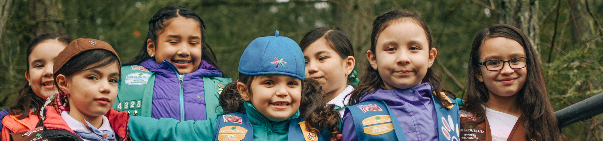  A group of Girl Scouts wearing blue and brown vests gathers in a forest and poses arm-in-arm smiling. 