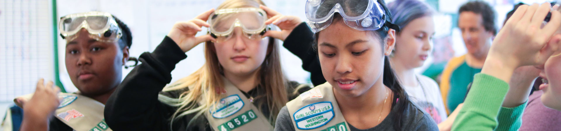  Three Girl Scouts wearing tan vests are seen putting on lab goggles. 