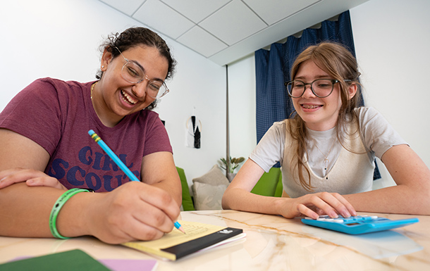 Two Girl Scouts working at a desk. One is writing on a notepad, the other is using a calculator.