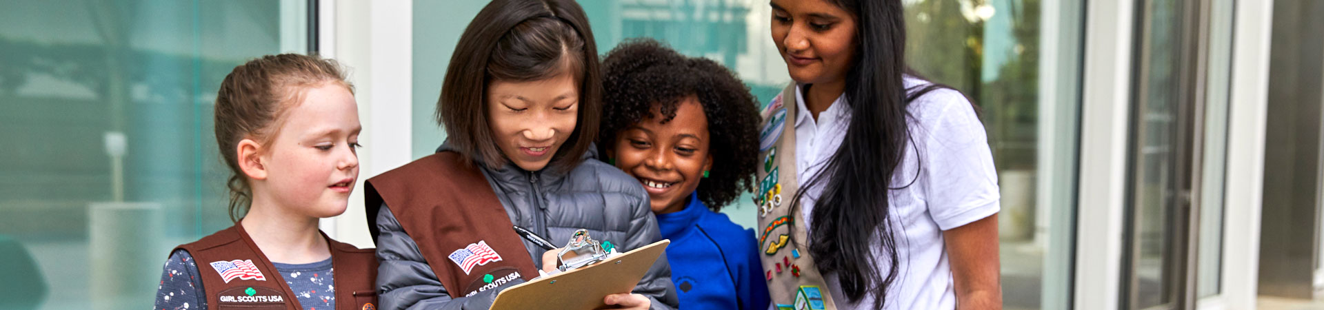  Four Girl Scouts stand behind a table full of cookie boxes, one holding a clipboard. 