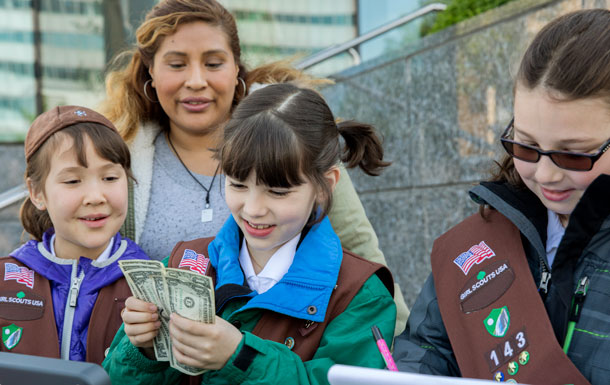 Three Girl Scouts outdoors counting money and taking notes. An adult volunteer supervises.