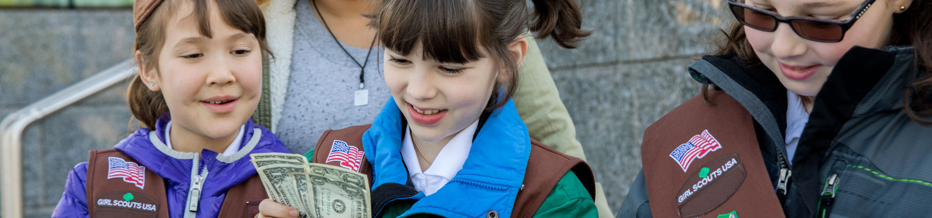  Three Girl Scouts in brown vests and sashes decorated with pins and badges stand at a booth counting money and taking notes. 