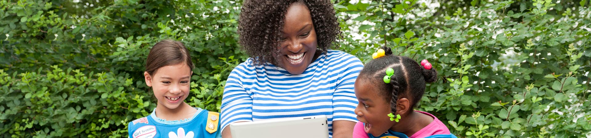  An adult volunteer sits between two Daisies wearing blue vests, holding an iPad for them to watch, 