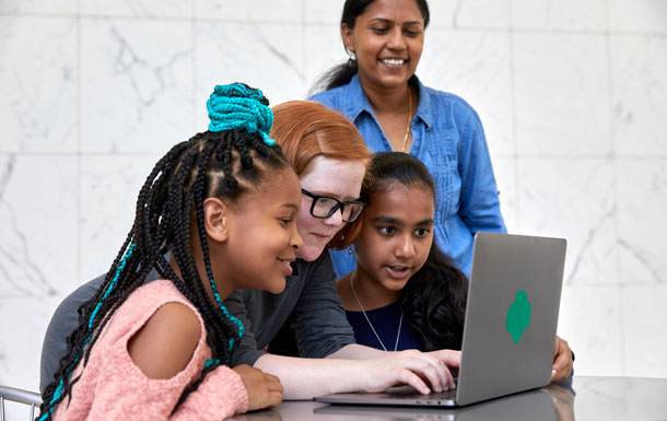 Three Girl Scouts examine an open gray laptop with a green Trefoil on the back with an adult volunteer wearing a jean shirt standing behind them.