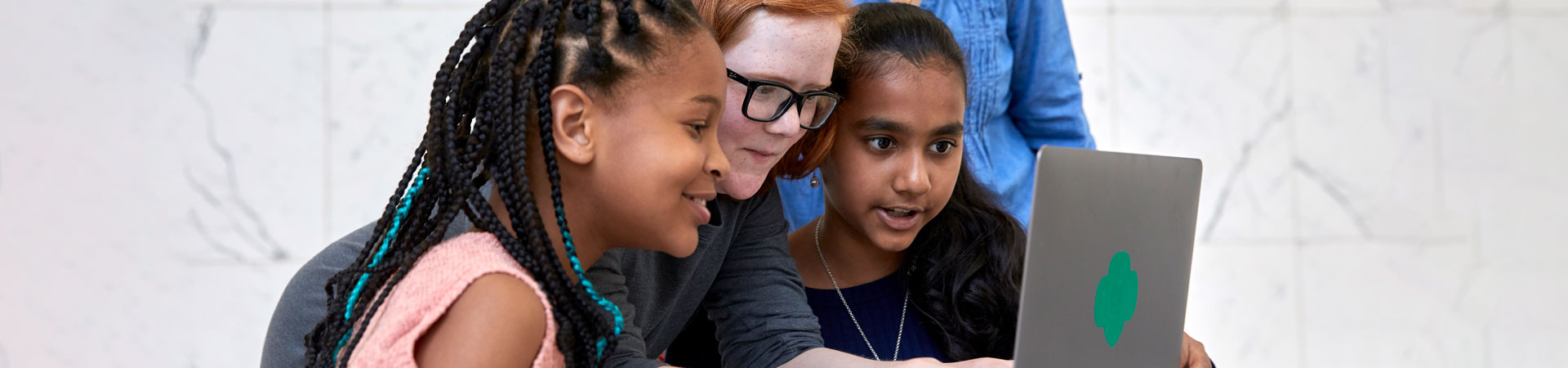  Three Girl Scouts examine an open gray laptop with a green Trefoil on the back with an adult volunteer wearing a jean shirt standing behind them. 