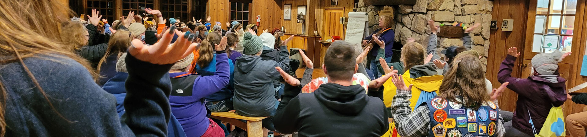  A group of volunteers gathers inside a camp lodge, sitting on wood benches with their backs to the camera. 