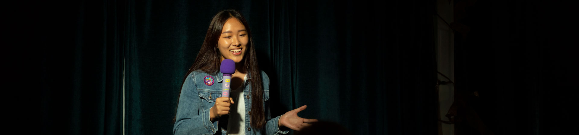  A speaker stands in front of a black background wearing a jean jacket and holding a microphone 