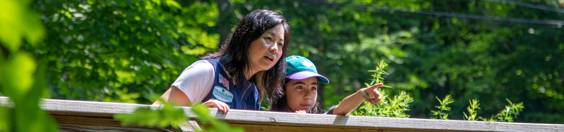  An adult volunteer stands outside on a wooden bridge next to a youth member pointing at something. 