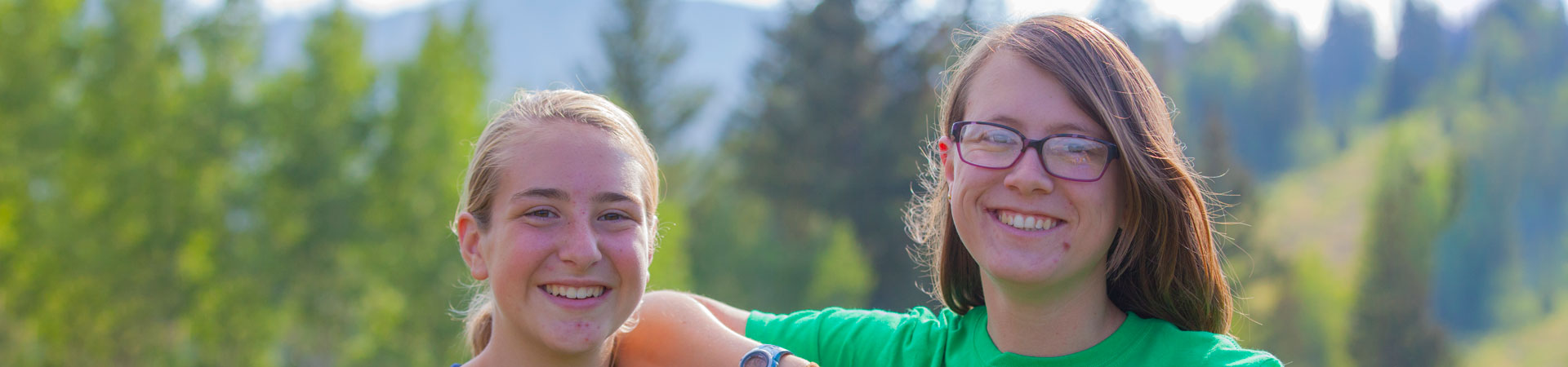  Two Girl Scouts wearing blue and green shirts are seen standing in front of green trees. 