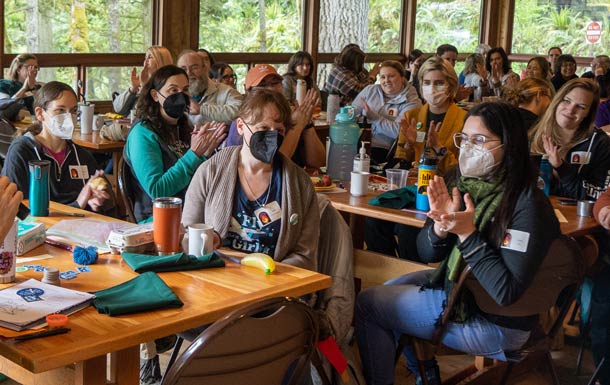 A group of staff and volunteers gathers in a camp lodge, sitting at wood tables and wearing masks.