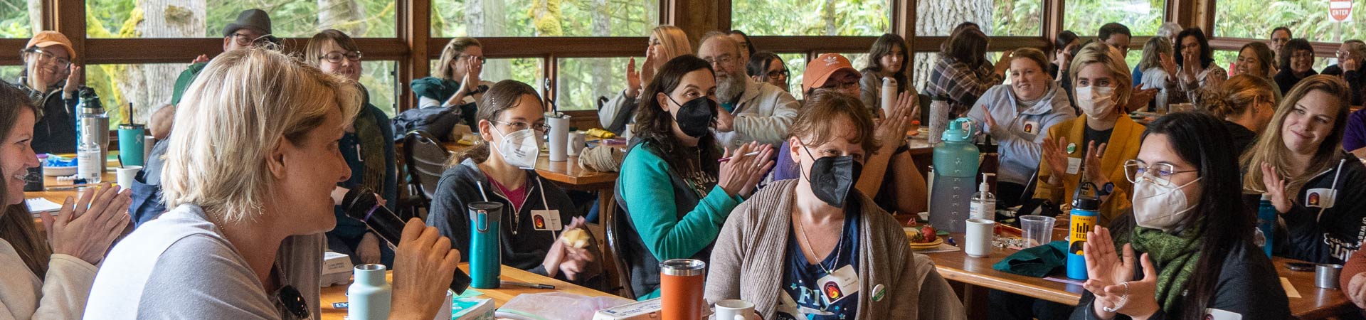  A group of staff and volunteers gathers in a camp lodge, sitting at wood tables and wearing masks. 