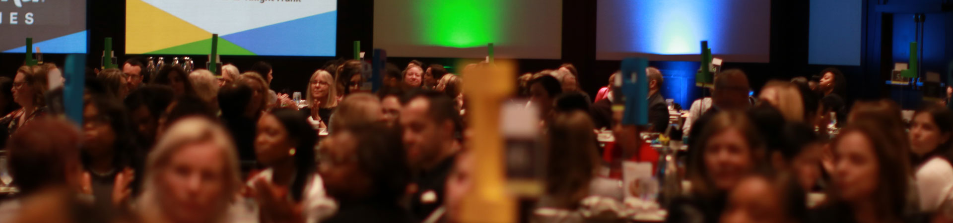  An indoor event filled with blurry attendees sitting at tables and a projector screen in the background. 