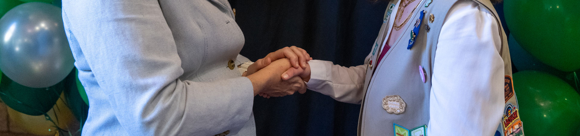  A Girl Scout shaking hands with one of Girl Scouts of Western Washington's community partners. There is a dark-blue stage curtain in the background. 