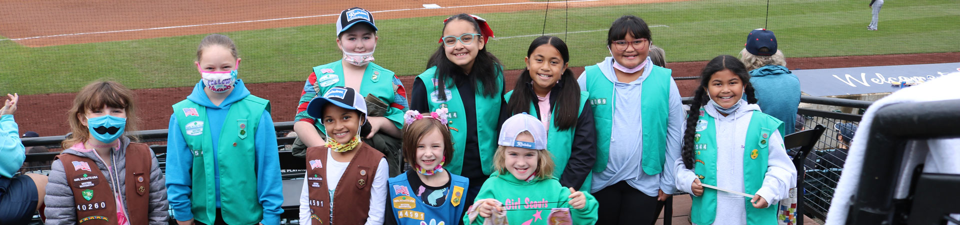  A group of Girl Scouts wearing brown, green, and blue vests poses outside in front of a baseball field. 