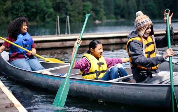 A group of three Girl Scouts paddles a canoe on a lake with a wood dock and green trees in the background.
