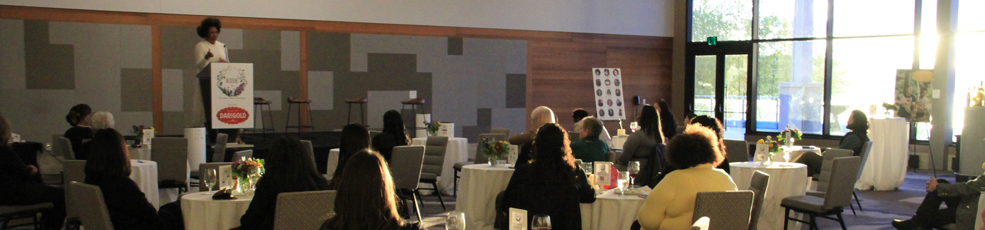  A speaker stands on a stage behind a podium during an indoor event with attendees seated at tables with white tablecloths. 