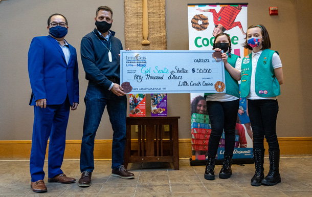 Two men in blue suits and wearing face masks hold an enlarged check with two Girl Scouts in green vests and wearing masks.