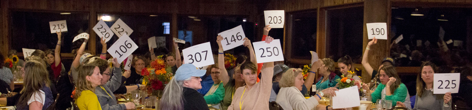  A group gathers in a camp lodge and sites at tables to participate in an auction where they raise a white piece of paper with a black number to bid. 