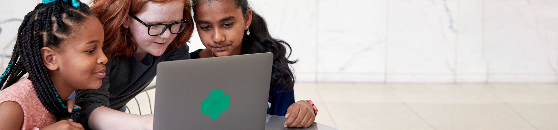  Three Girl Scouts examine an open gray laptop with a green Trefoil on the back. 