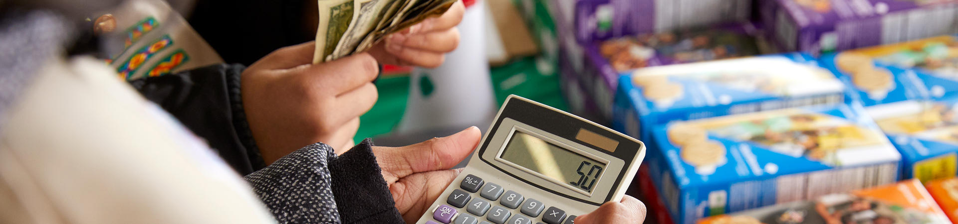  The hands of two Girl Scouts are seen in front of a stack of cookies, one holding a calculator and one counting cash. 