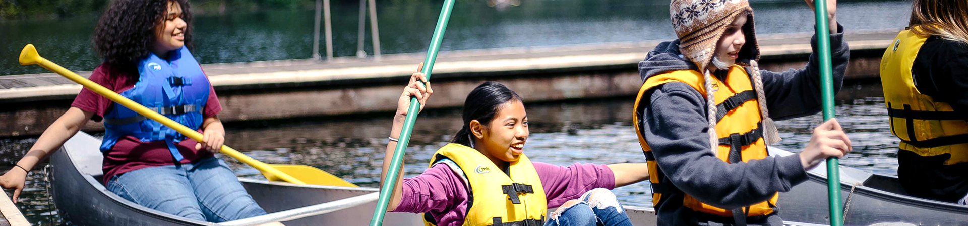  Three Girl Scouts paddle in a canoe on a lake with a dock in the background. 
