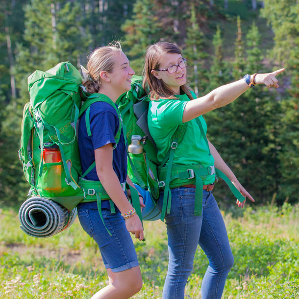 Two Girl Scouts wearing large green backpacks are standing in a grassy field with trees in the background. One Girl Scout points to something ahead on the trail.