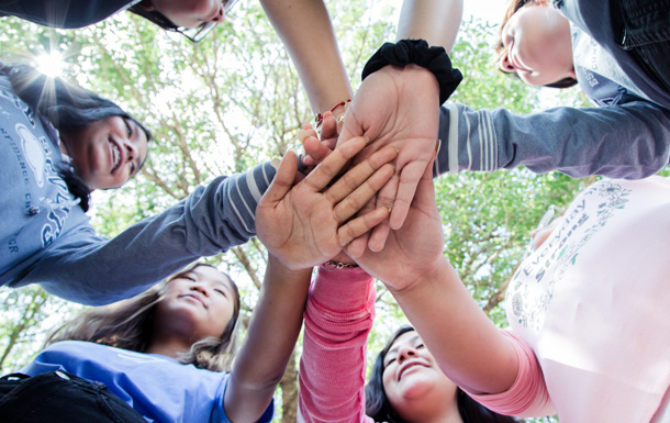 A group of Girl Scouts standing outside with trees visible over heads each put one of their hands into the middle of the circle.